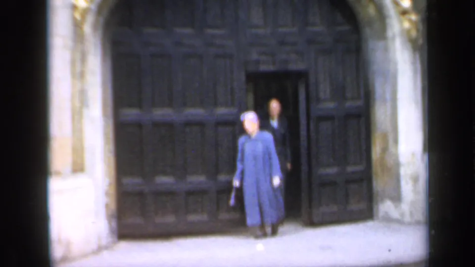 A person in a blue coat stands or walks near the large dark wooden cathedral door, with another visitor partly visible behind.