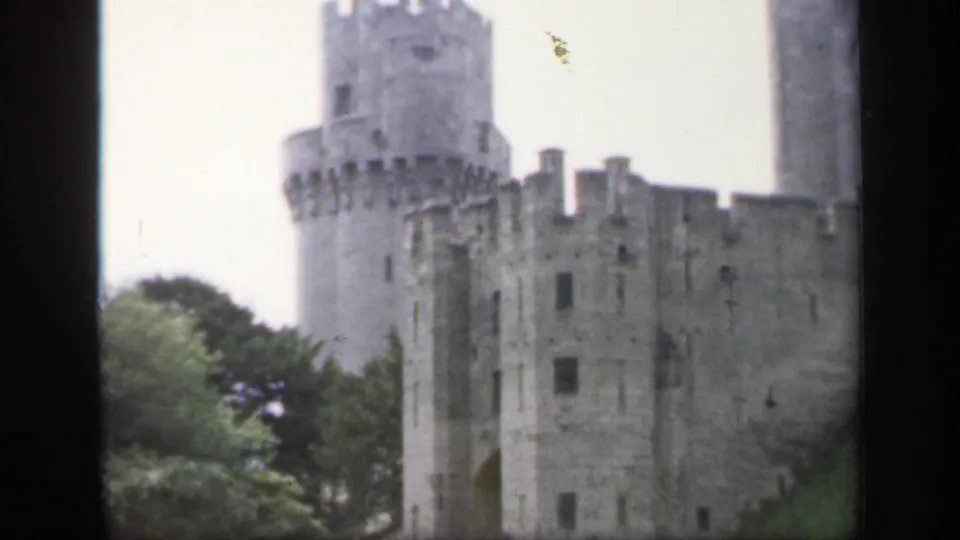 1949 England 8mm frame showing pale stone castle towers, crenellated walls, trees, and overcast sky from a visitor viewpoint.