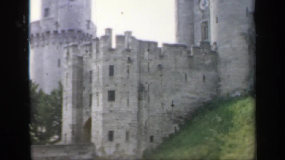 1949 England 8mm frame of a stone castle wall, tower, arched opening, trees, and grassy bank in soft handheld focus.