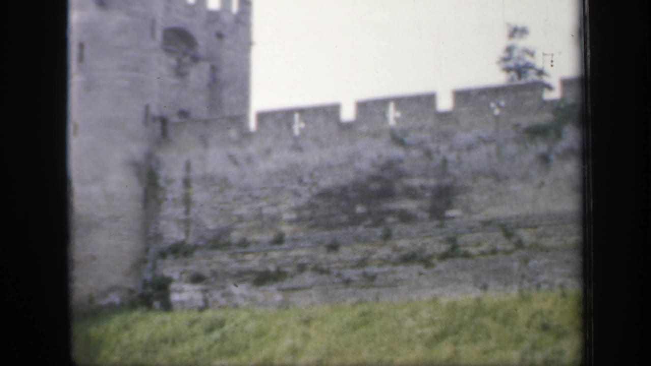 1949 England 8mm frame showing a gray stone castle wall, partial tower, grass, and dark film-scan edges.