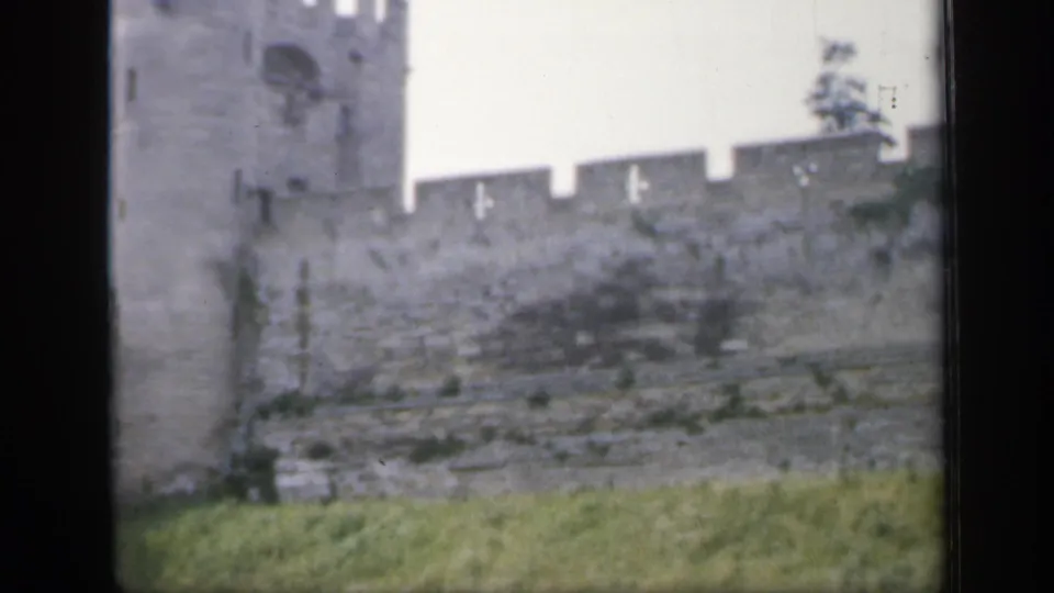 1949 England 8mm frame showing a gray stone castle wall, partial tower, grass, and dark film-scan edges.