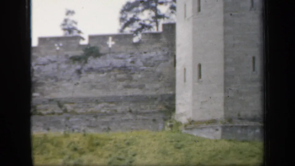 1949 England 8mm frame with a tall stone tower, narrow openings, crenellated wall, grass, and trees in the background.