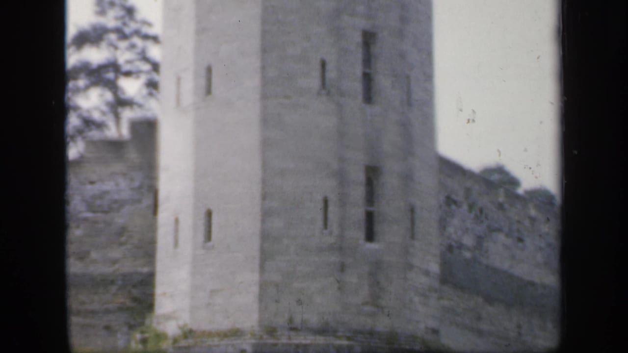 1949 England 8mm frame showing a tall pale stone tower, narrow vertical openings, adjoining wall, and trees in soft focus.