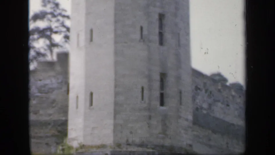 1949 England 8mm frame showing a tall pale stone tower, narrow vertical openings, adjoining wall, and trees in soft focus.