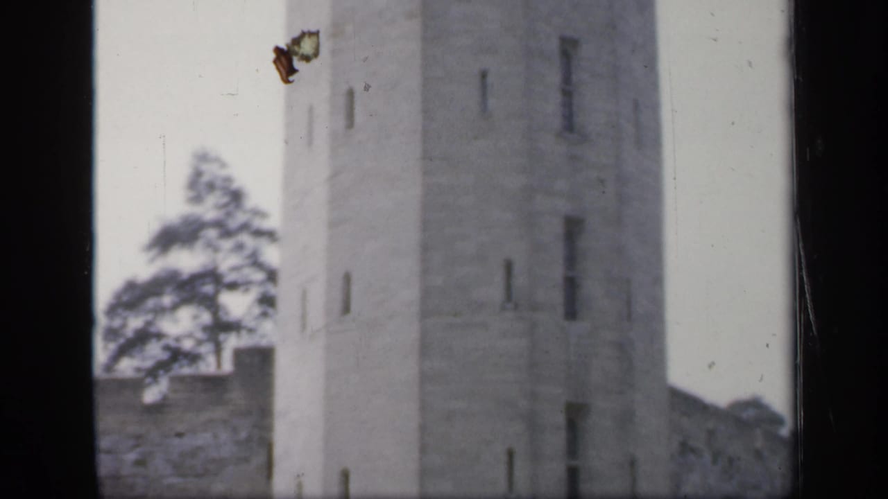 1949 England 8mm frame with a tall weathered stone tower, slit-like windows, adjoining wall, trees, and pale sky.