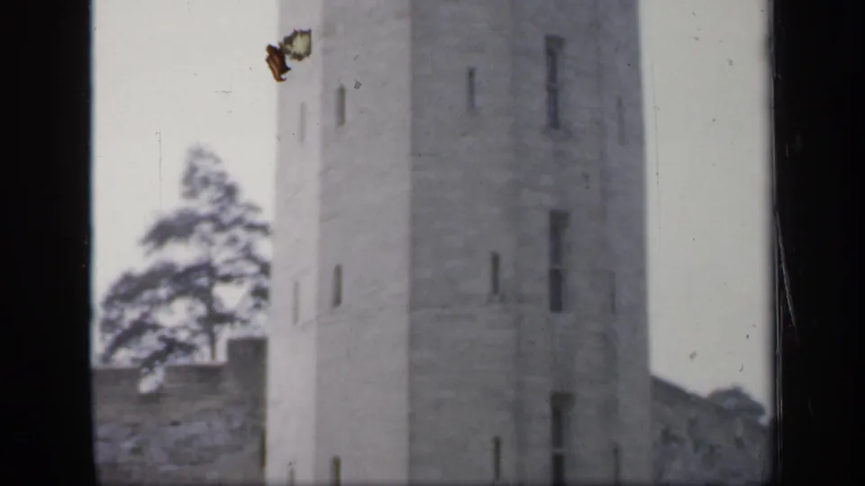 1949 England 8mm frame with a tall weathered stone tower, slit-like windows, adjoining wall, trees, and pale sky.