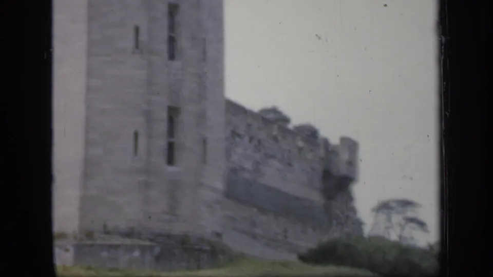 1949 England 8mm frame showing a rectangular stone tower, crenellated wall, and sloped grass below the castle exterior.