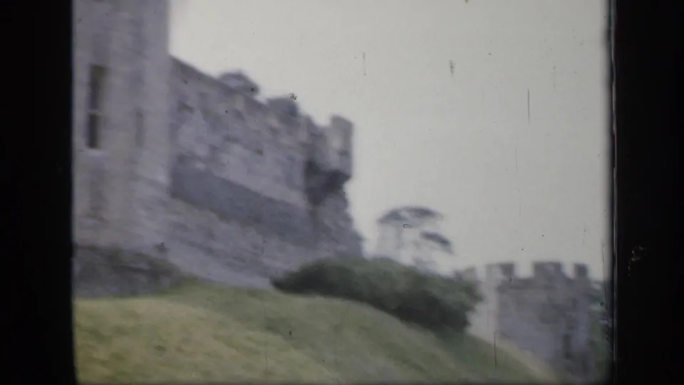 1949 England 8mm frame showing castle stonework along a grassed slope with a distant crenellated section and trees nearby.