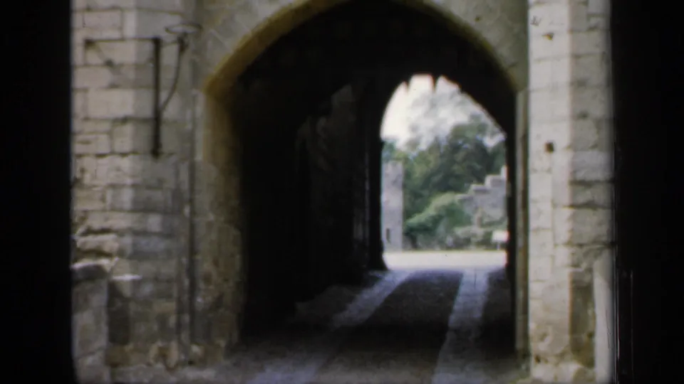 1949 England 8mm frame showing a shadowed stone castle archway with a paved passage and greenery visible outside.