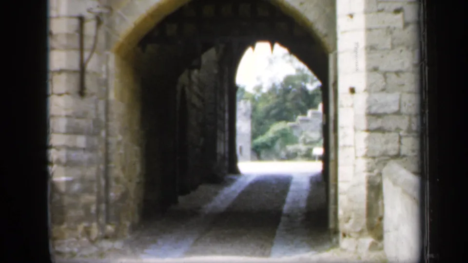 1949 England 8mm view through a castle passage, showing arched stonework, iron gate sections, and greenery beyond.