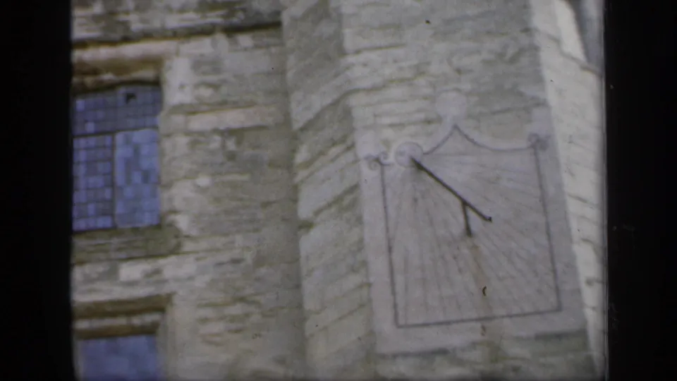 1949 England 8mm view of a blue-tinted leaded window beside a sundial on an old stone facade.