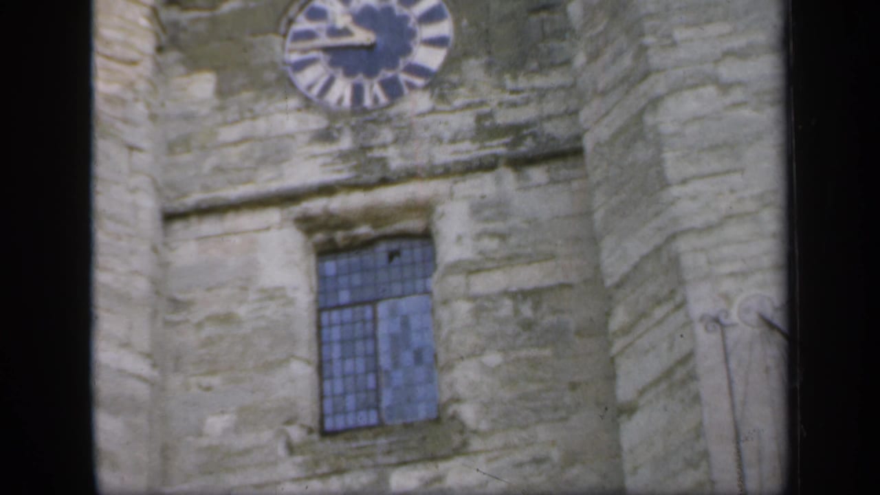 1949 England home-movie frame showing weathered castle stonework, a round clock face, and a blue-tinted leaded window.