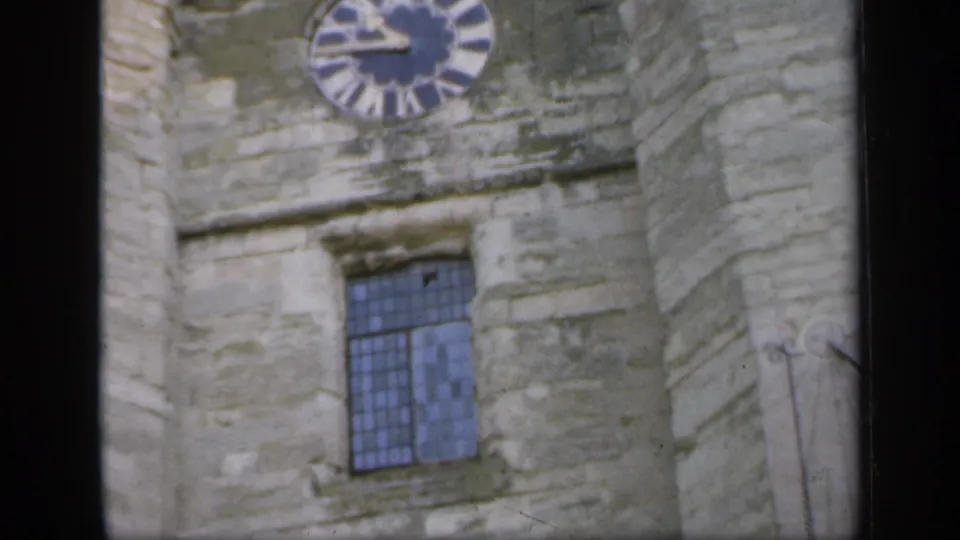 1949 England home-movie frame showing weathered castle stonework, a round clock face, and a blue-tinted leaded window.