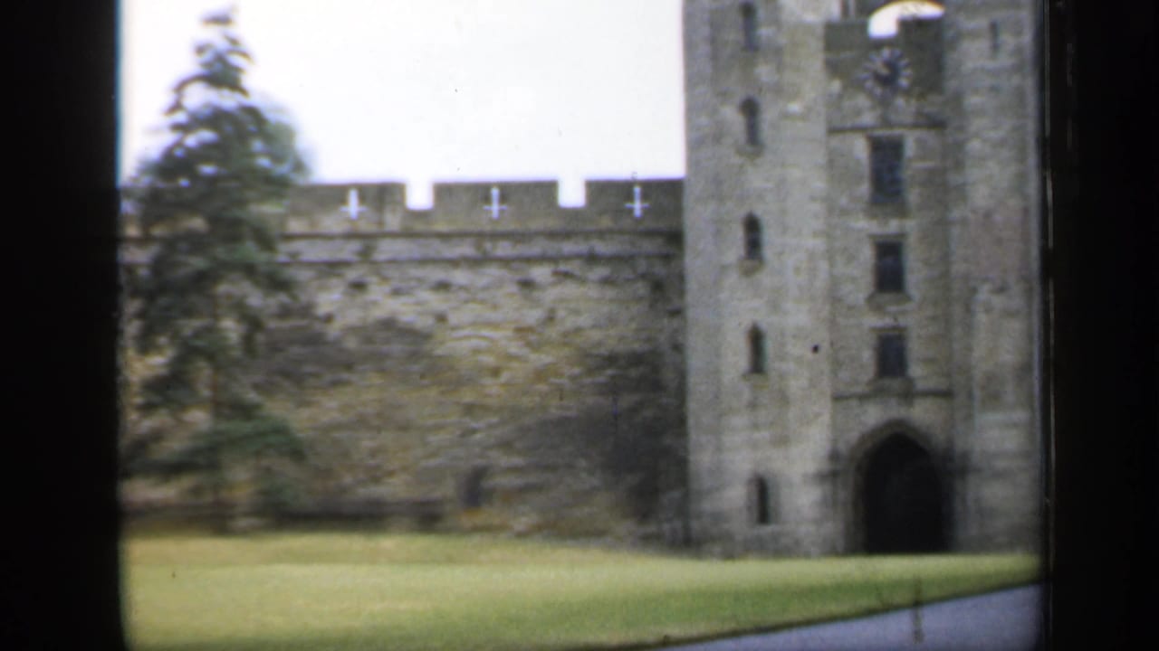1949 England castle exterior with a crenellated wall, tower gateway, green lawn, and mature trees in soft 8mm focus.