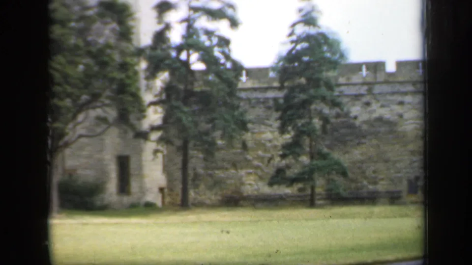1949 England castle view showing crenellated stone wall, mature trees, open lawn, and part of a tower in soft home-movie color.