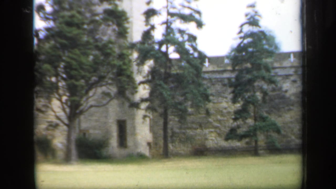 1949 England 8mm frame of a stone castle wall, lawn, and tall evergreen trees seen from a visitor viewpoint.