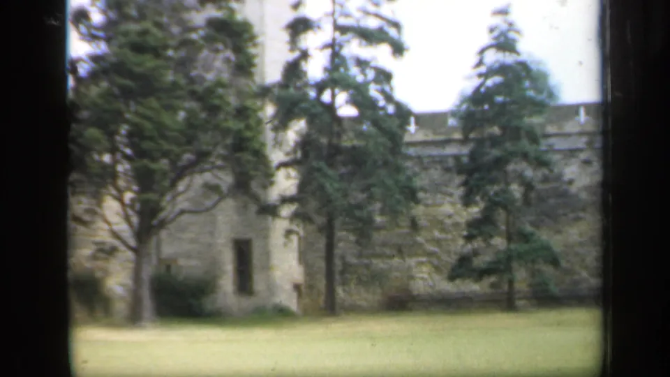 1949 England 8mm frame of a stone castle wall, lawn, and tall evergreen trees seen from a visitor viewpoint.