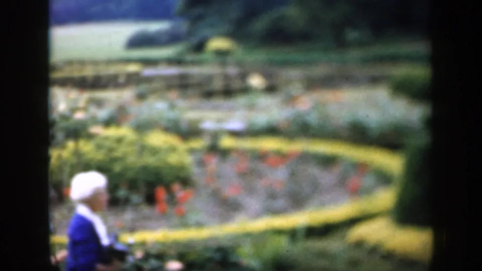 1949 England garden frame with soft-focus beds, clipped hedges, paths, and a partial blue-and-white shape at the lower left edge.