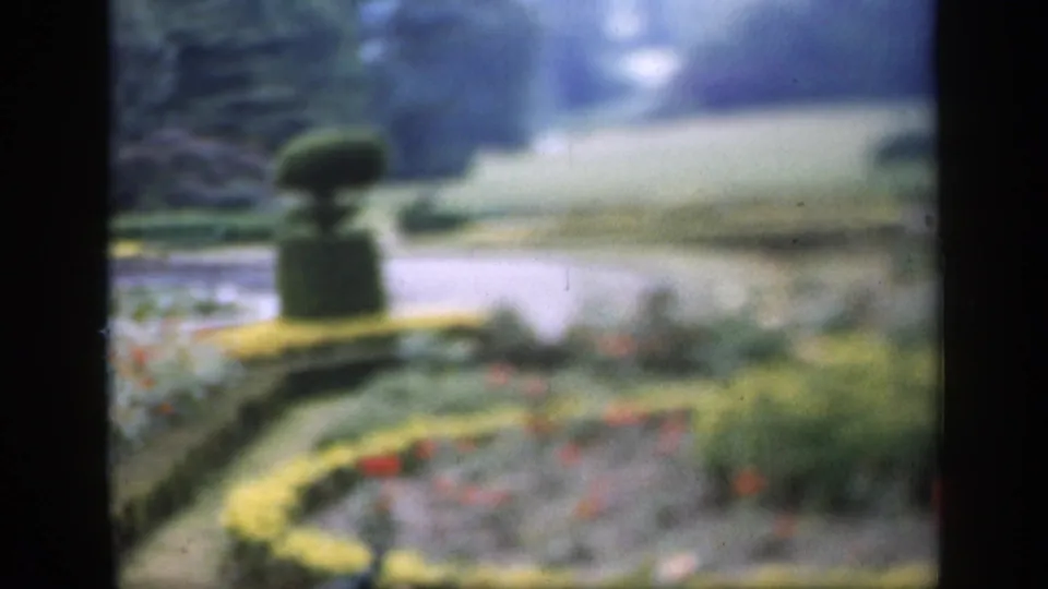1949 England 8mm frame showing clipped garden borders, colorful flowers, a curved path, and central topiary in soft focus.