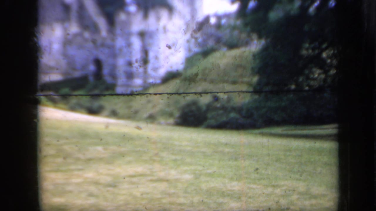 1949 England 8mm frame showing a historic stone castle wall beyond a sloped lawn, path, shrubs, and trees.