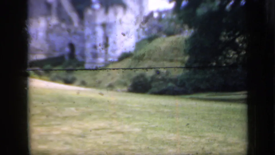 1949 England 8mm frame showing a historic stone castle wall beyond a sloped lawn, path, shrubs, and trees.