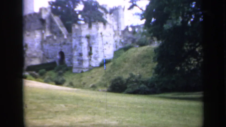 1949 England 8mm frame showing castle stonework, a grassy bank, open lawn, shrubs, and dark tree cover.