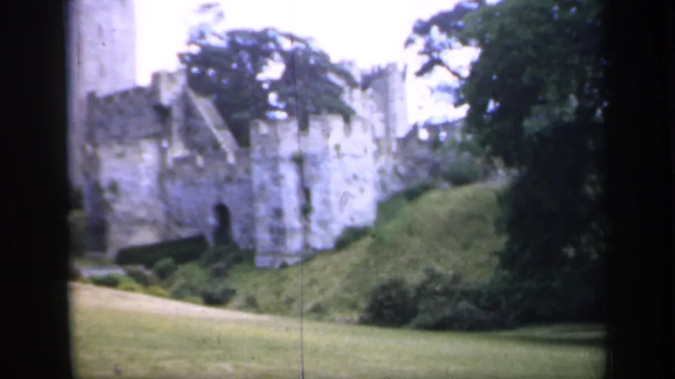 1949 England 8mm frame showing a historic stone castle wall beside a grassy slope, with trees and soft home-movie texture.