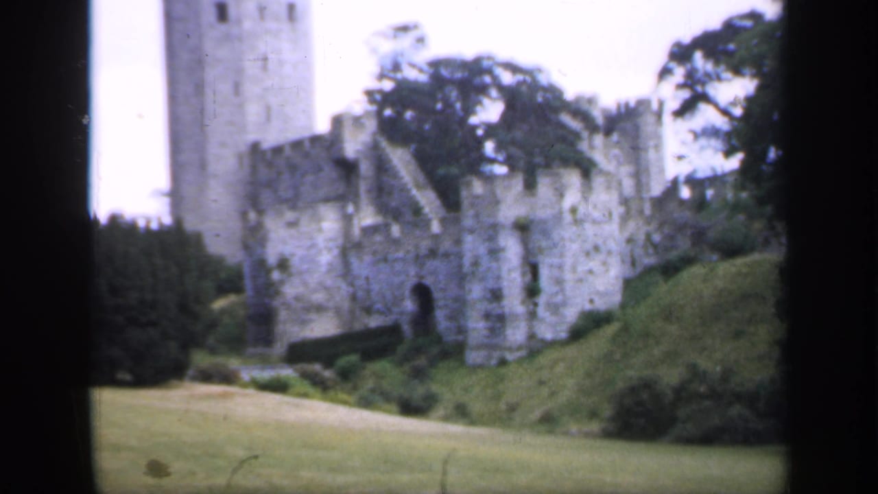 1949 England home-movie frame of a stone castle tower and wall line seen across lawns with trees in the background.