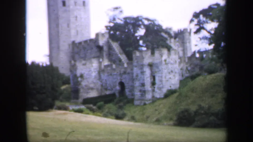 1949 England home-movie frame of a stone castle tower and wall line seen across lawns with trees in the background.