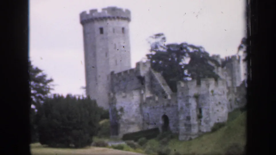 1949 England 8mm frame showing the round tower, adjoining castle walls, dark tree line, and green embankment.