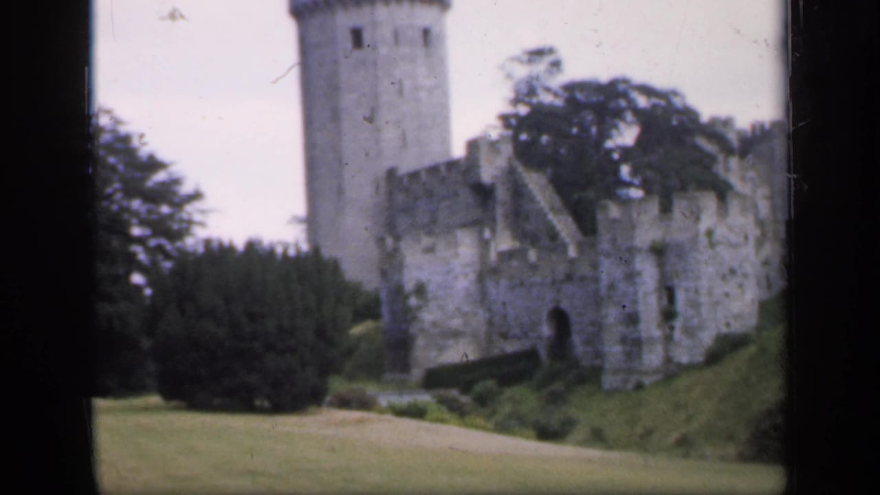 1949 England 8mm frame showing a stone castle, round tower, arched entry, lawn, and trees from a visitor viewpoint.
