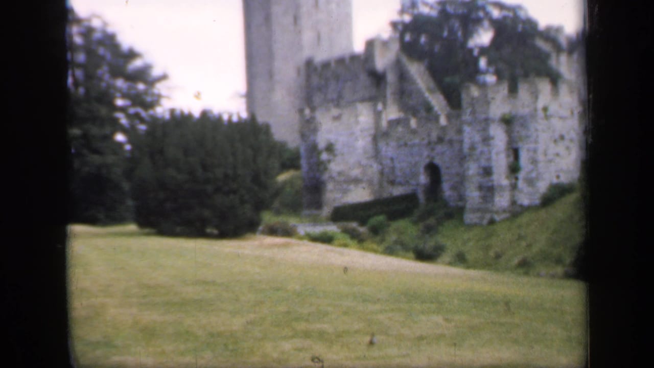 1949 England home-movie frame with a castle tower, weathered stone wall, lawn, hedges, and mature trees in soft color.