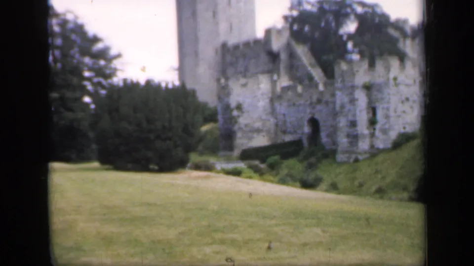 1949 England home-movie frame with a castle tower, weathered stone wall, lawn, hedges, and mature trees in soft color.