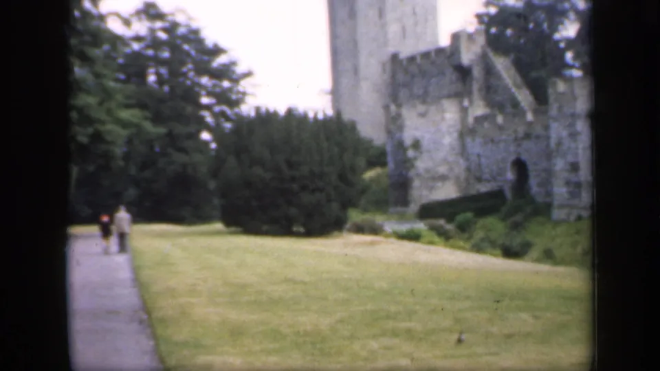1949 England 8mm frame showing castle grounds, lawn, trees, and two distant visitors walking along a paved path.