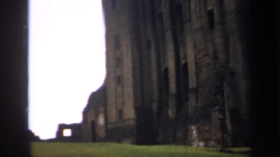 1949 England home-movie frame looking along grass toward stone castle ruins, with dark doorway-like edges.