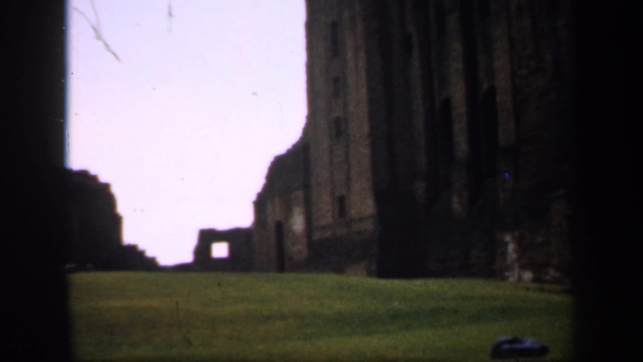 1949 England 8mm frame showing a grassy lawn leading toward weathered stone ruins, partly blocked by dark foreground masonry.