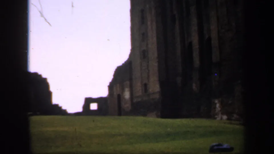1949 England 8mm frame showing a grassy lawn leading toward weathered stone ruins, partly blocked by dark foreground masonry.