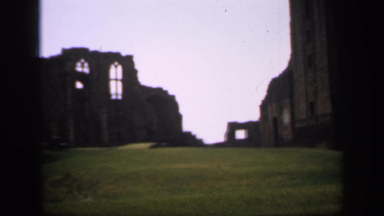 1949 England 8mm frame centered on weathered stone ruins with large Gothic-style window openings above a sloped lawn.