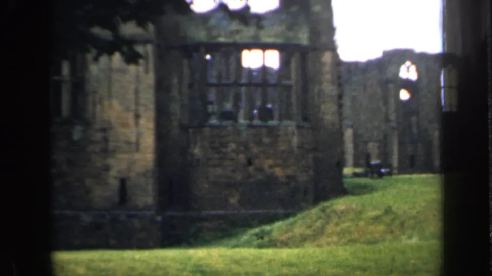 Closer 8mm view of historic-looking English stone ruins with window openings, rough masonry, and a sloping green lawn.