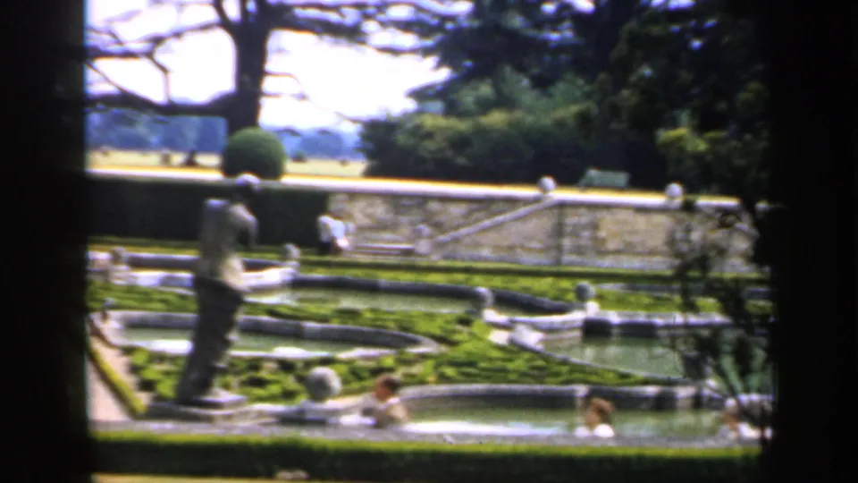 1949 England home-movie view of a formal garden with low fountains, clipped hedges, benches, and distant visitors on the paths.