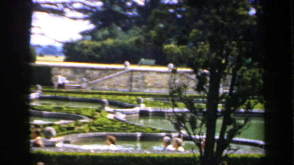 1949 England 8mm view of a formal garden with stone pools, clipped hedges, trees, and distant visitors behind dark foreground framing.