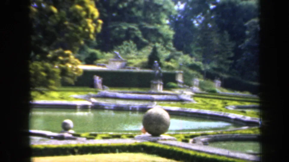 1949 England 8mm frame of a formal garden with green pools, a round stone ornament, hedges, statues, and distant visitors.