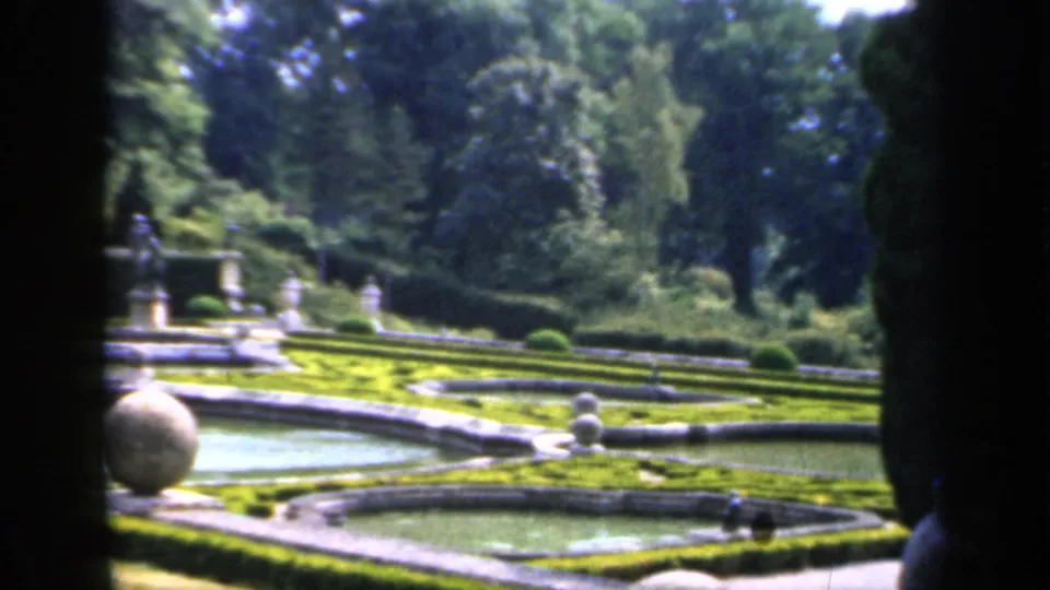 1949 England home-movie view across stone-edged garden pools toward clipped hedges, statues, dense trees, and distant visitors.