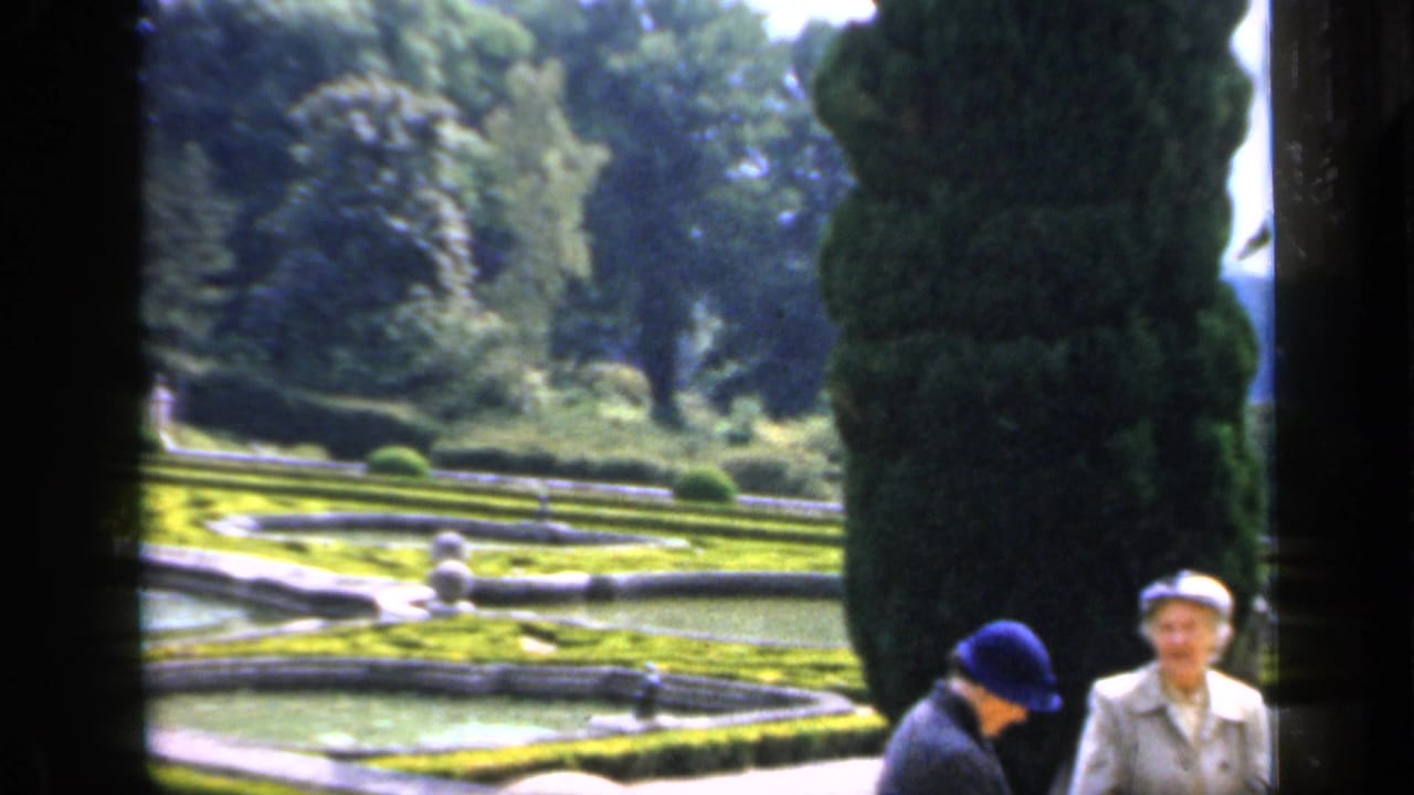 In England archival 8mm footage, two adult women stand near a reflecting pool, clipped hedges, statues, and tall evergreens in a formal garden.