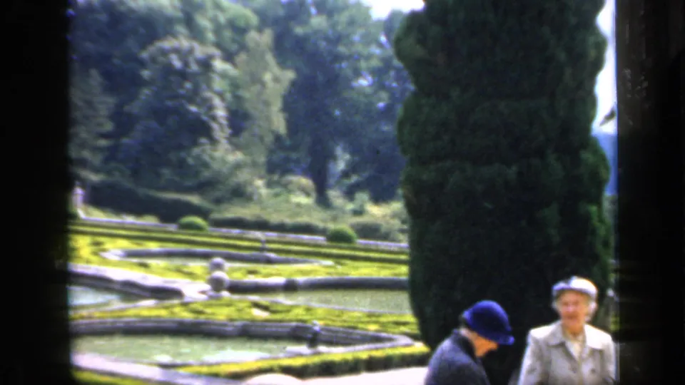 In England archival 8mm footage, two adult women stand near a reflecting pool, clipped hedges, statues, and tall evergreens in a formal garden.
