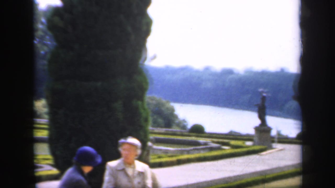 Two adult women in a formal English garden appear between dark 8mm frame edges, with a path, lake, clipped hedges, and statue visible behind them.