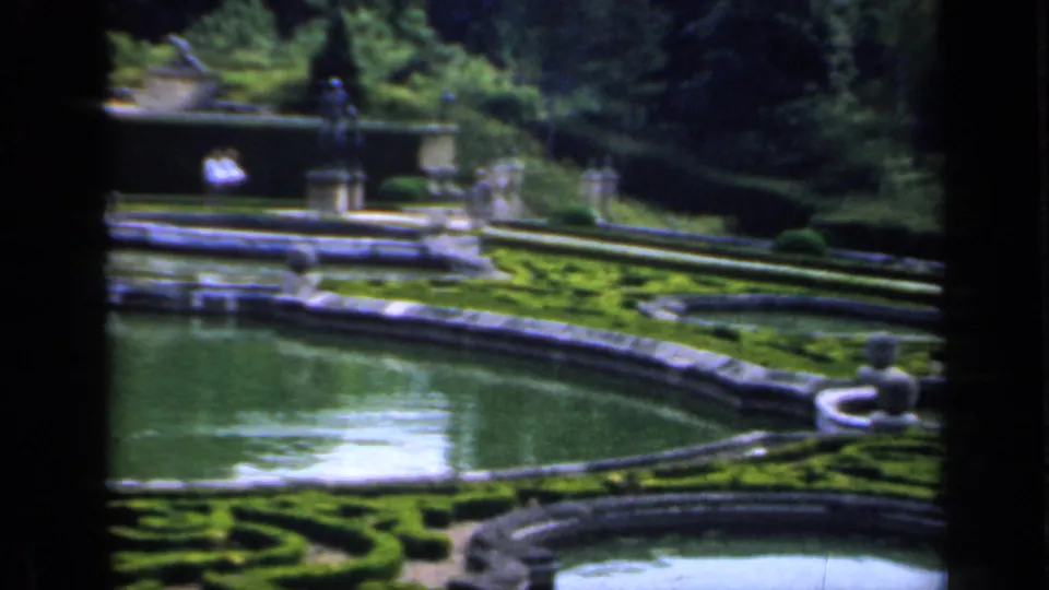 A formal garden scene shows reflecting pools, clipped hedges, ornamental stonework, statues, and dense trees in soft 8mm color.