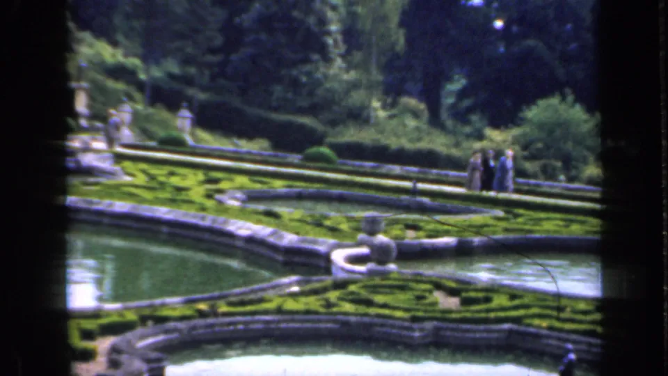Adult visitors walk in the distance through a formal English garden with reflecting pools, clipped hedges, stone edging, and ornamental statues.