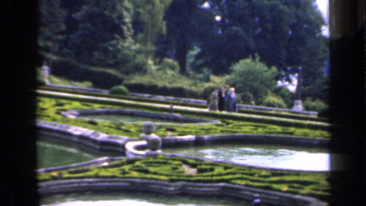 1949 England home-movie frame showing a formal garden with terraced hedges, a pale fountain basin, mature trees, and distant visitors.