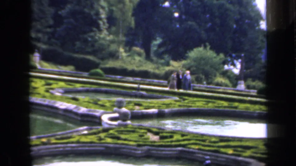 1949 England home-movie frame showing a formal garden with terraced hedges, a pale fountain basin, mature trees, and distant visitors.
